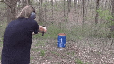 a man is shooting a gun in the woods near a blue barrel