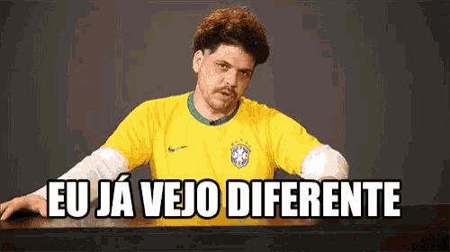 a man in a yellow shirt is sitting at a table with the words eu ja vejo diferente above him .
