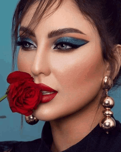 a woman with blue eyeshadow and red lipstick is holding a red rose in her mouth