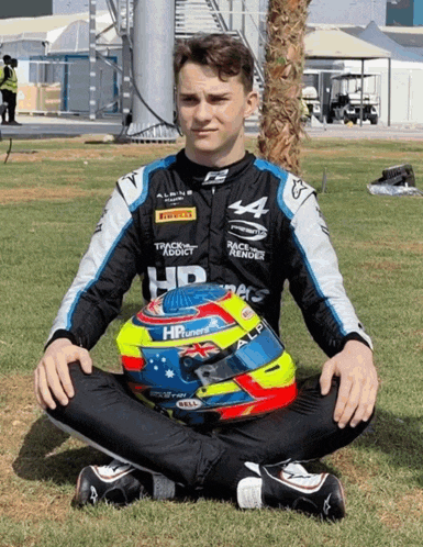 a man in a race suit sits on the grass with his helmet