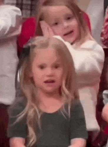 two little girls are sitting next to each other in a room . one of the girls is making a funny face .