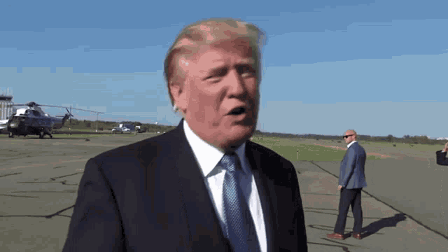 a man in a suit and tie is standing on a runway with a helicopter in the background