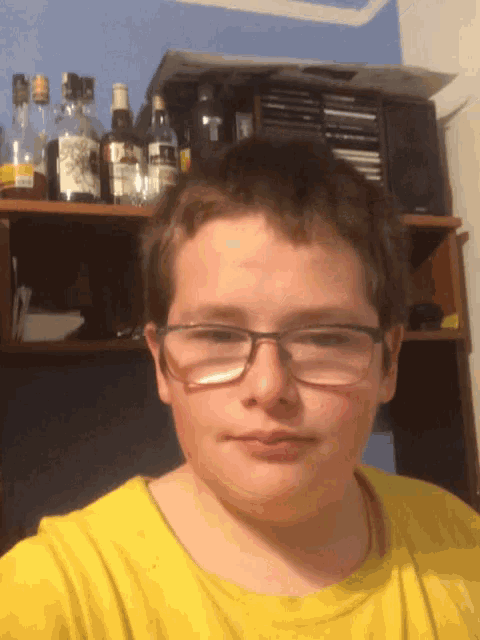 a boy wearing glasses and a yellow shirt stands in front of a shelf full of bottles
