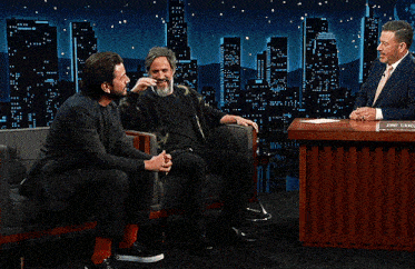 a man with a beard is sitting next to another man in a suit