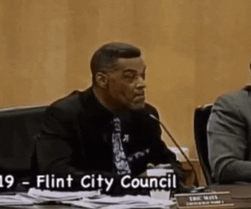 a man in a suit and tie is speaking into a microphone with the words flint city council written below him