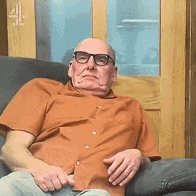 an elderly man wearing glasses is sitting in a chair making a funny face .