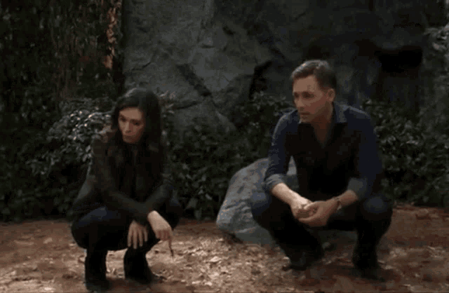 a man and a woman are squatting in the dirt near a rock .