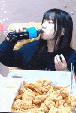 a girl drinking from a bottle while sitting at a table with fried chicken and french fries