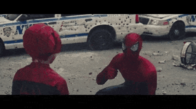two children dressed in spiderman costumes are standing in front of a police car covered in bullet holes .
