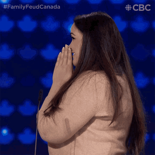 a woman covering her mouth with her hands while standing in front of a microphone with the hashtag familyfeud canada on the bottom right