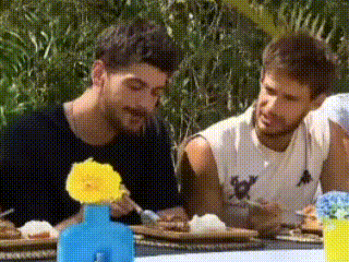 two men are sitting at a table eating food with a flower in a vase .