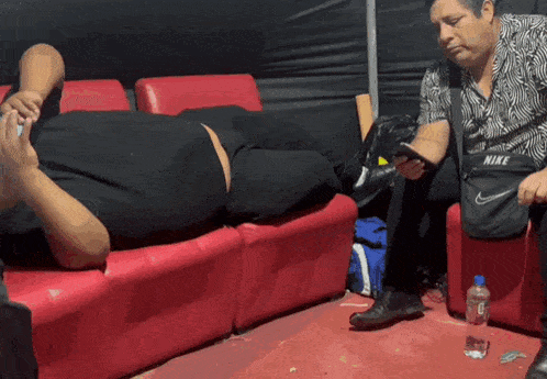 a man wearing a nike purse sits next to a man laying on a couch