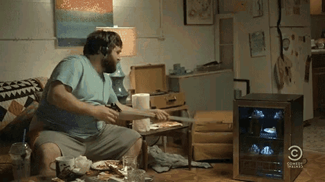 a man is sitting on a couch eating pizza next to a comedy central fridge