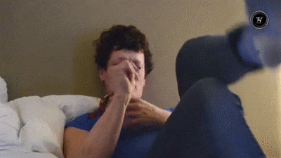 a man in a blue shirt is laying on a bed covering his nose with his hand .