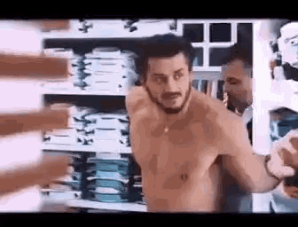 a man without a shirt is standing in front of a stack of shirts in a store .