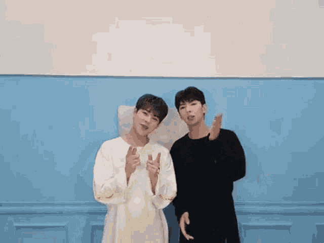 two young men are standing next to each other in front of a blue wall and making a peace sign .