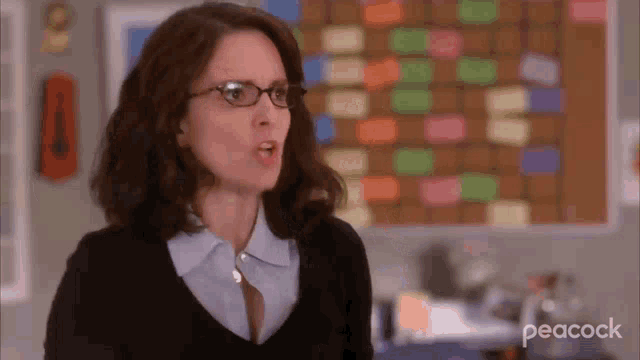 a woman wearing glasses and a black sweater is making a funny face in front of a bulletin board .