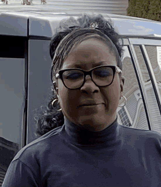 a woman wearing glasses and a blue turtleneck is standing in front of a car .