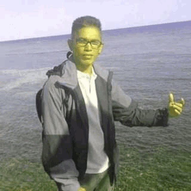 a young man wearing glasses and a jacket is standing on a beach giving a thumbs up .