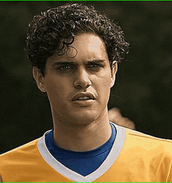 a young man with curly hair is wearing a yellow shirt and a blue t-shirt .