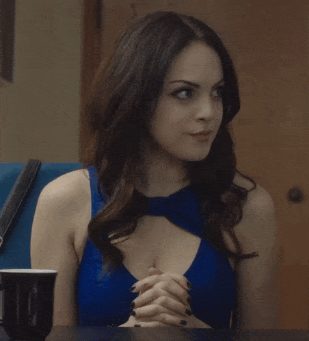 a woman in a blue dress with a bow around her neck is sitting at a table with her hands folded