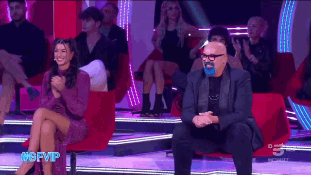 a man with a blue mask on his face sits next to a woman in a purple dress applauding .