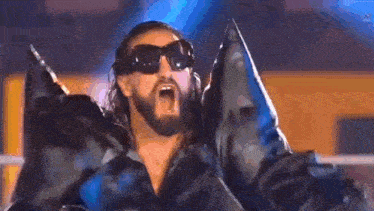 a man with a beard wearing sunglasses and a black jacket is standing in a ring with his mouth open .