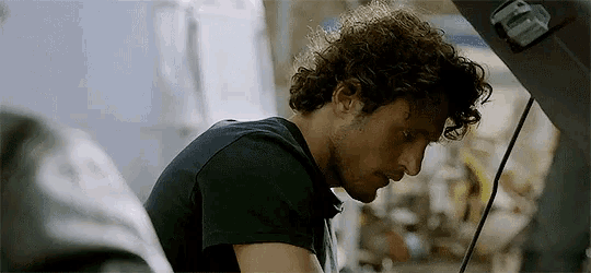 a man with curly hair looks under the hood of his car