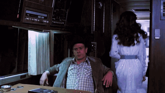 a man in a plaid shirt sits at a table while a woman in a white dress walks past him