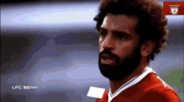 a soccer player with a beard is wearing a red shirt and looking at the camera .