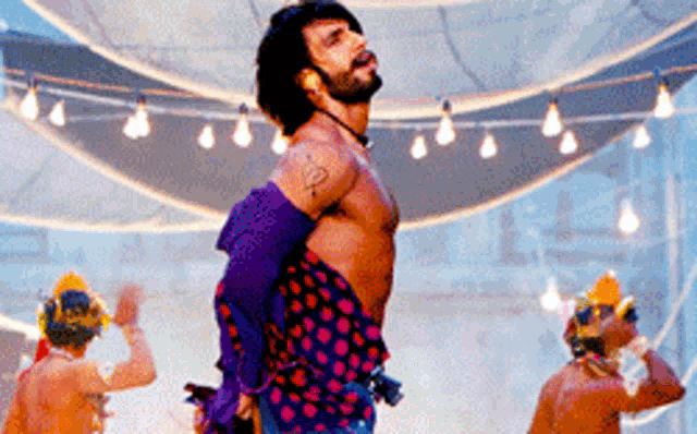 a shirtless man in a polka dot shirt is dancing in a room with a string of lights hanging from the ceiling .
