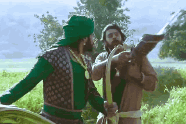 two men are standing in a field one is holding a bow and arrow and the other is holding a gun