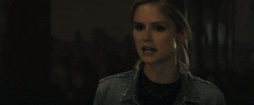 a woman wearing a denim jacket and a black shirt is looking at the camera in a dark room .