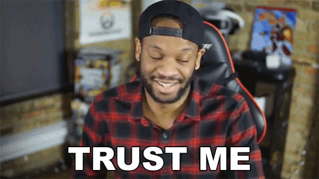 a man wearing a hat and a plaid shirt is sitting in a chair and smiling with the words trust me behind him .