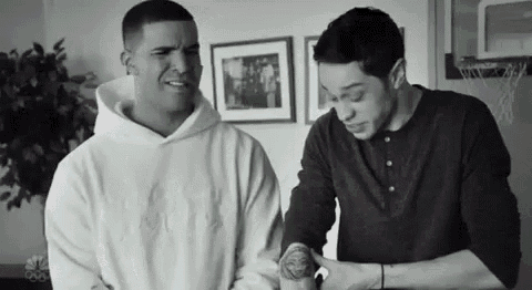 a black and white photo of two men standing next to each other . one of the men is holding a tattoo on his arm .