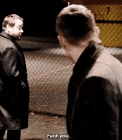 two men are standing next to each other in front of a chain link fence and talking .
