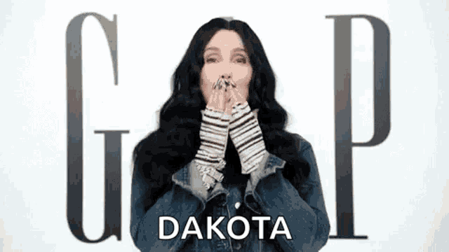 a woman in a denim jacket is covering her mouth with her hands in front of the gap logo .