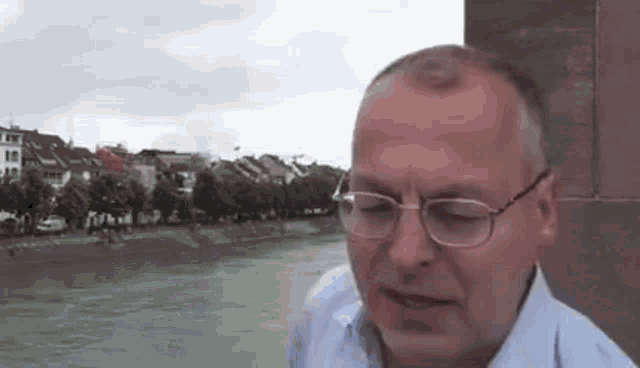 a man wearing glasses is standing next to a river and talking .
