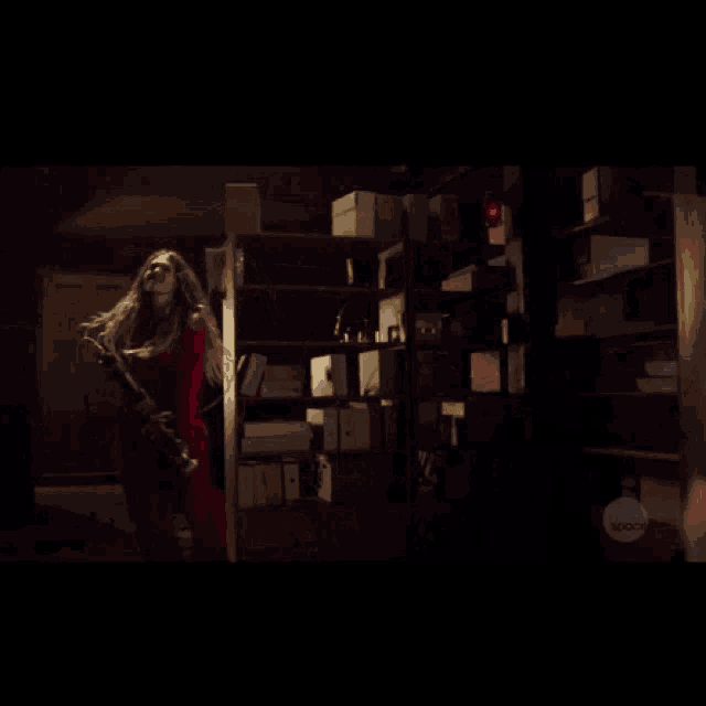 a woman in a red coat is holding a gun in a dark room with boxes on shelves