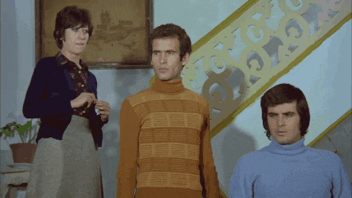 a man in an orange sweater stands in front of a woman and a man in a blue shirt