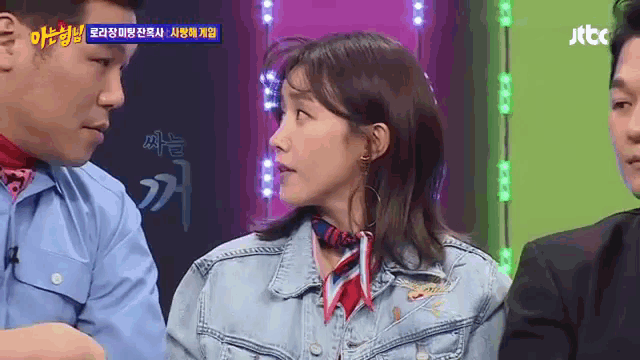 a woman in a denim jacket with a red scarf around her neck talks to a man in a suit on a tv show