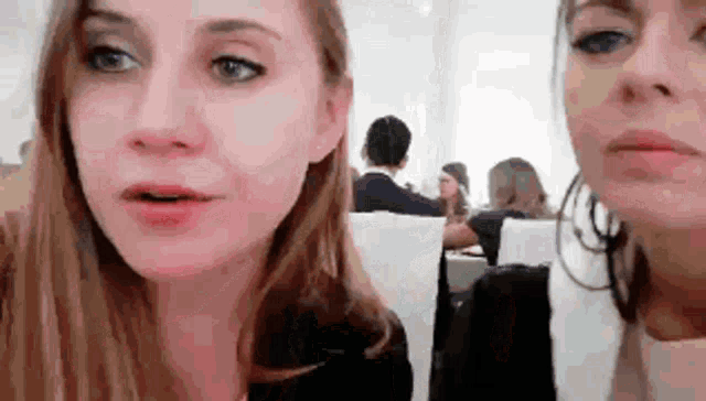 two women are sitting next to each other in a room with people sitting at tables .