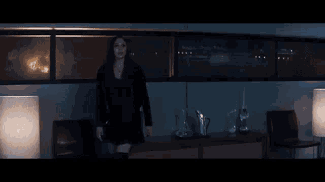 a woman in a black jacket is standing in a dark room with lamps and chairs .