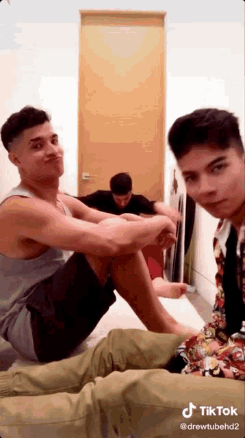 a group of young men are sitting on a bed and one of them is wearing a tank top .