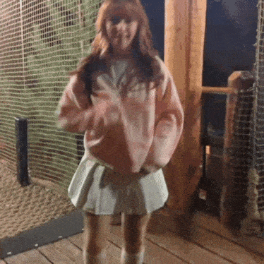 a girl in a pink sweater and white pleated skirt is dancing on a deck .