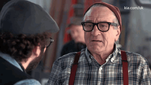 a man in a plaid shirt and suspenders talks to another man in a kia.com/uk advertisement