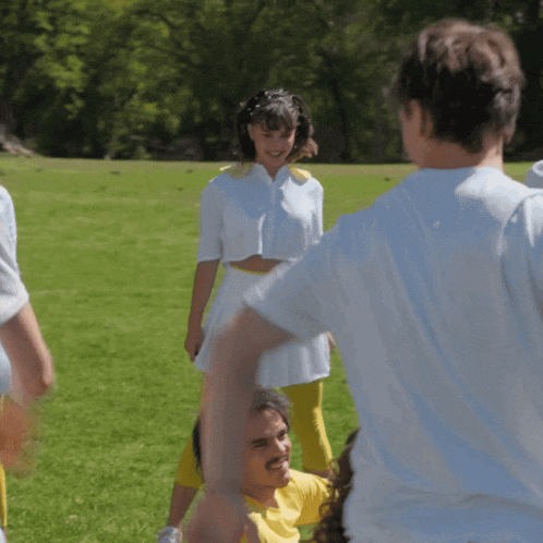 a woman in a white crop top and yellow pants stands in a field with other people