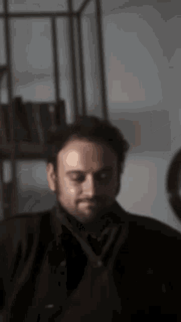 a man with a beard is sitting in front of a bookshelf and looking down .