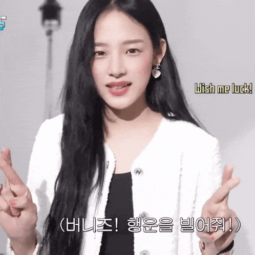 a woman with long black hair is wearing a white jacket and making a wish me luck gesture