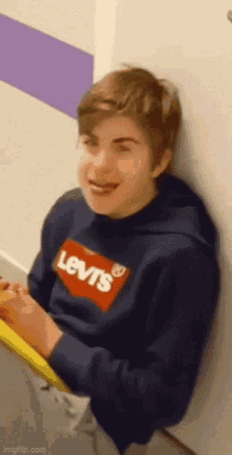 a young boy wearing a blue levi 's sweatshirt is leaning against a wall and laughing .